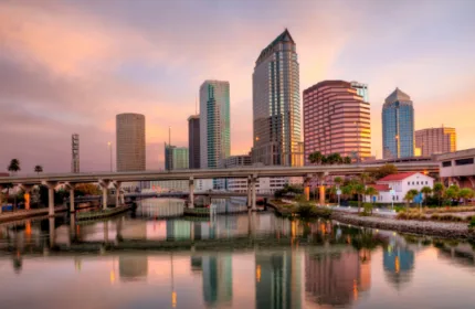 Downtown Tampa skyline at sunset with modern high-rise buildings reflecting on the water, viewed from across the river with a bridge in the foreground.