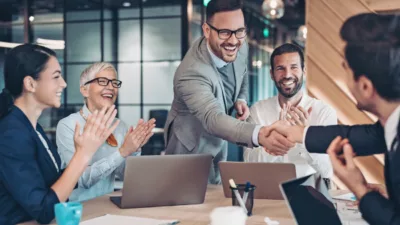 Smiling professionals in business attire celebrate a successful meeting in a modern office, with two shaking hands across the table as colleagues applaud. Image represents blog covering, what is an executive?