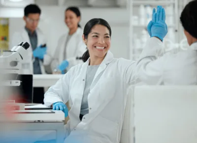 A team of happy scientists in lab coats celebrate with high fives in a bright, modern laboratory. This image captures collaboration, teamwork and scientific breakthroughs—perfect for showcasing a positive research environment.