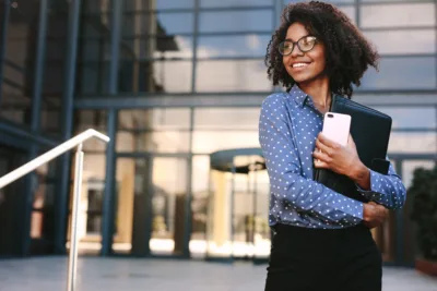 Business professional standing outside a modern office building holding a smartphone and portfolio.