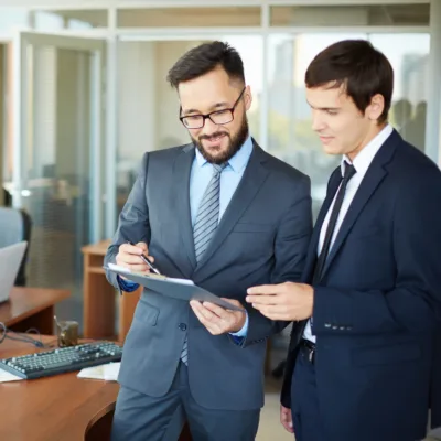 Two professionals are talking in a bright office hallway, one holding a tablet, representing an optimized contractor hiring process and improved workforce collaboration in a pharmaceutical organization.