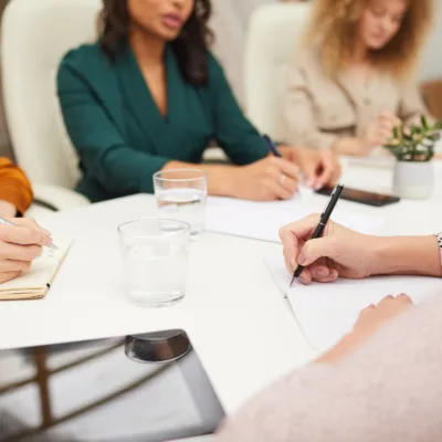 Close-up of hands writing notes at a table during a meeting with others in the background, representing collaboration supporting accounts payable conversion projects.