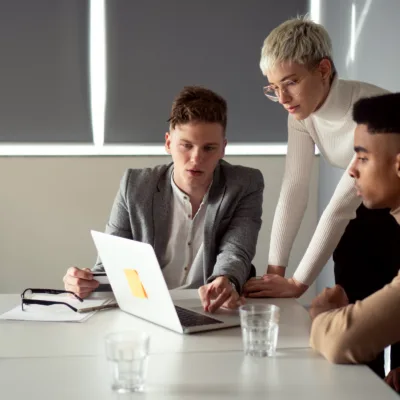 Three professionals working together at a table reviewing information on a laptop, representing collaboration across onsite and remote teams to meet urgent pharmacovigilance timelines.