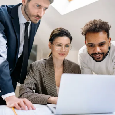 Three professionals gathered around a laptop reviewing information together, representing collaboration and detailed analysis for a pharmacovigilance auditing project.