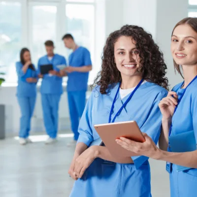 Two healthcare professionals in scrubs holding a tablet with a care team in the background, representing quickly scaling HEDIS nurse staffing and supporting high volume healthcare projects.