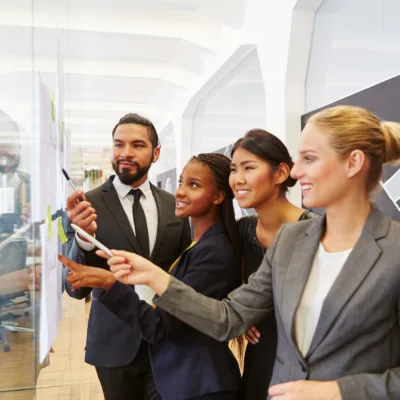 Four professionals gathered around a glass board reviewing notes and pointing to documents, representing building organizational structure and developing standard operating procedures.