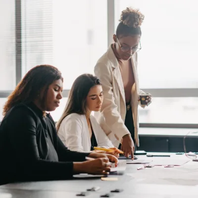 Three professionals collaborate at a desk reviewing documents and pointing to notes, representing teamwork, guidance, and hands-on problem solving in a modern office setting.