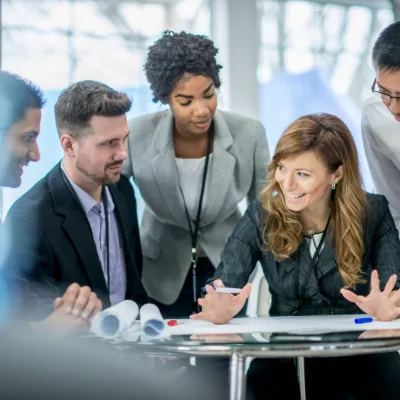 A group of professionals gather around a table reviewing documents and discussing ideas collaboratively in a modern office setting.