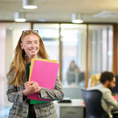 A professional stands in a modern office holding colorful folders and a portfolio, representing organization, preparedness, and confidence in a workplace setting.