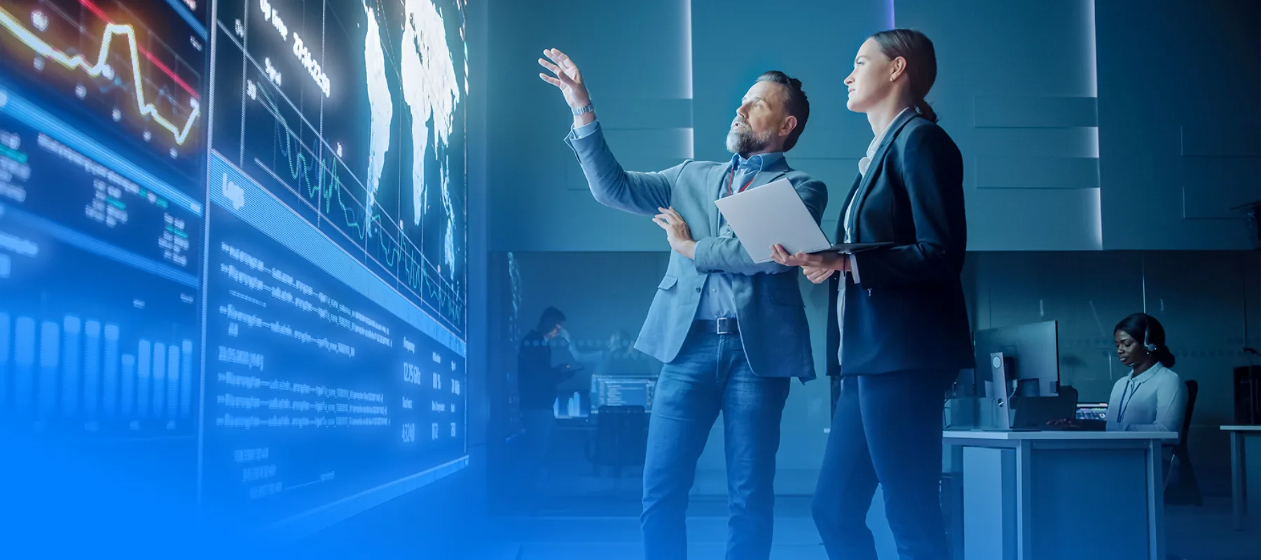 Colleagues in a highly technical workplace with digital screens covering a wall they are standing in front of. Man and woman talking with man pointing at the screens and woman looking on while holding a laptop. Behind them a woman wearing a headset and ID badge sits at a desk with multiple computer monitors.