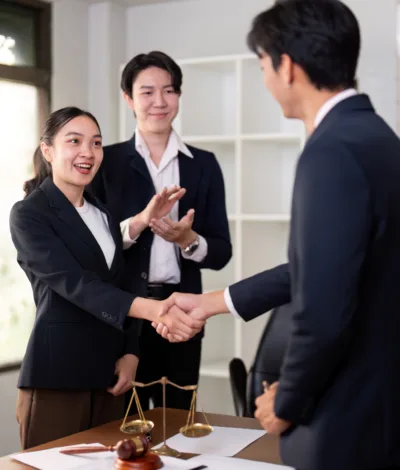 Legal professionals meeting and shaking hands in an office setting with case files and scales of justice on the table.