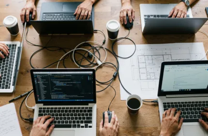 Laptops with code on the screen at a telecommunications business with ethernet cables and USB cords between them, people using external corded mouses.