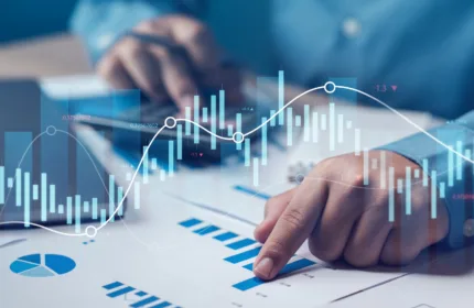 Close‑up of a person reviewing financial charts at a desk, pointing to data on printed graphs overlaid with digital stock and market indicators.
