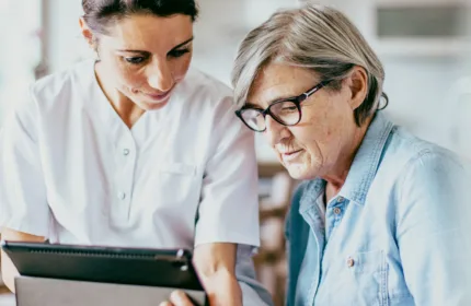 A healthcare professional sits beside an older adult, showing information on a digital tablet as they review it together in a home-like setting.
