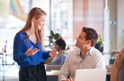 A woman standing and speaking to a colleague who is seated at a desk with a laptop in a bright, modern office setting.