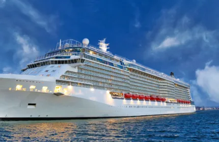 A large white cruise ship sails on calm blue water at dusk, illuminated by soft lighting against a dramatic sky filled with deep blue clouds.