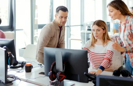 Engineers collaborating at computer workstations in a modern office, reviewing network systems and infrastructure displayed on multiple monitors.