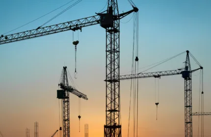 Several tall construction cranes silhouetted against an orange and blue sunset sky, with the sun low on the horizon behind an unfinished building.