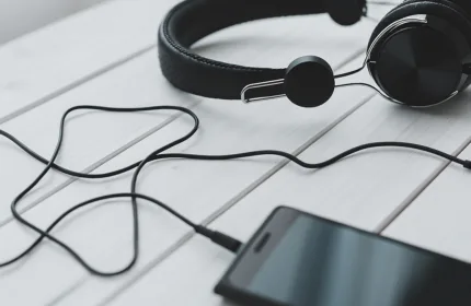 Headphones and a smartphone connected by a cable resting on a light wooden surface.