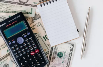 Calculator resting on scattered U.S. dollar bills next to a small spiral notebook and a silver pen on a white surface.