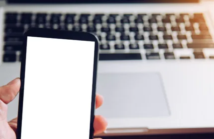Person holding a smartphone with a blank white screen in front of an open laptop on a wooden desk, representing modern electronic devices and digital technology.