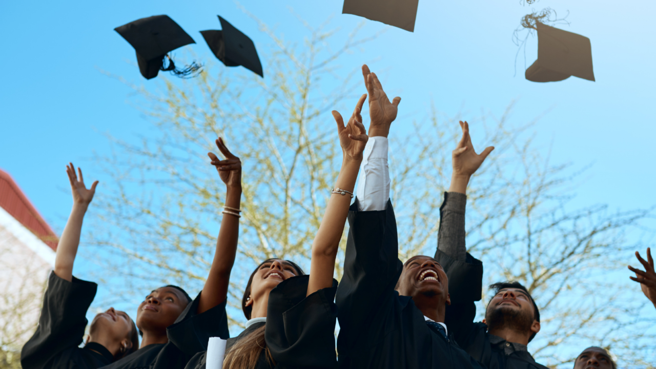 Graduates wearing black caps and gowns toss their mortarboards into the air outdoors, holding diplomas in hand. The scene represents academic achievement and celebration in an educational setting.