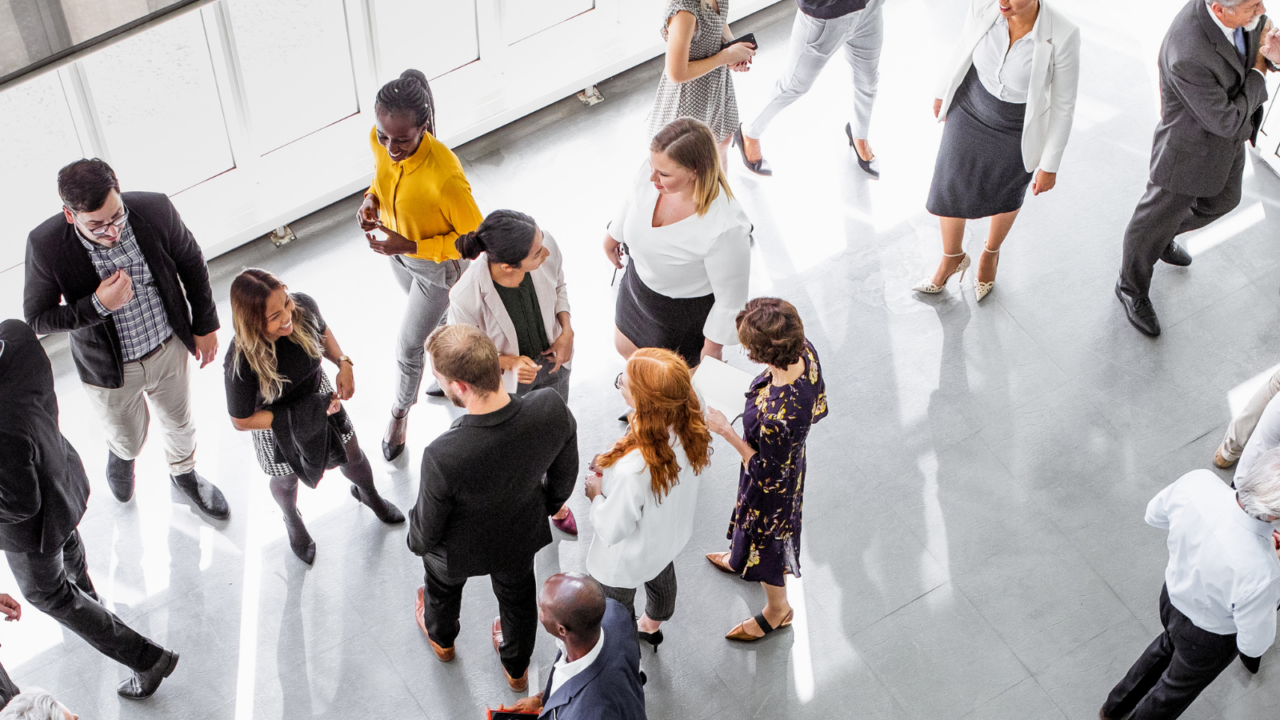A large group of professionals gathered in a bright, modern indoor space, engaging in conversations and networking. The setting includes people standing in small clusters, some holding drinks or tablets, representing a corporate event or business gathering.