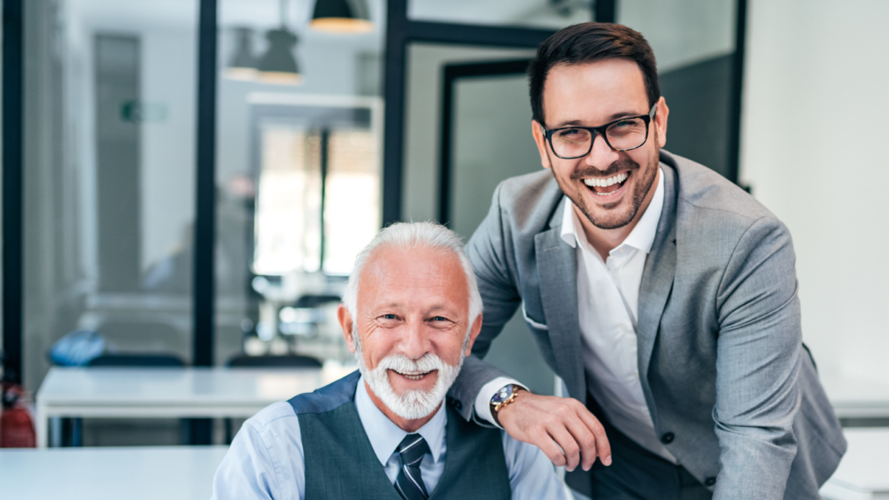 Two professionals in a modern office setting collaborate at a desk with a laptop. One person is seated working on the laptop while the other stands nearby, reviewing documents. The environment features glass walls and contemporary furniture, representing family office operations and financial management.