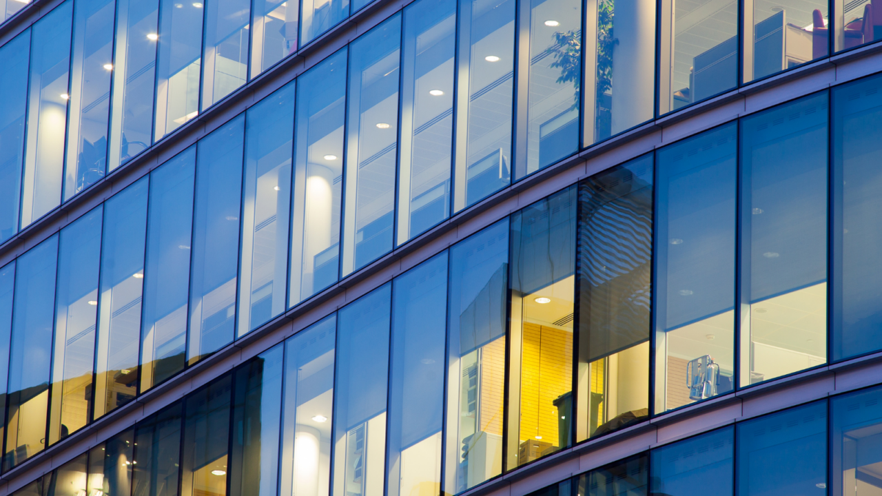Exterior view of a contemporary office building with large glass windows revealing multiple floors. Inside, well-lit workspaces, meeting areas, and modern furnishings are visible, showcasing a professional and collaborative office environment.