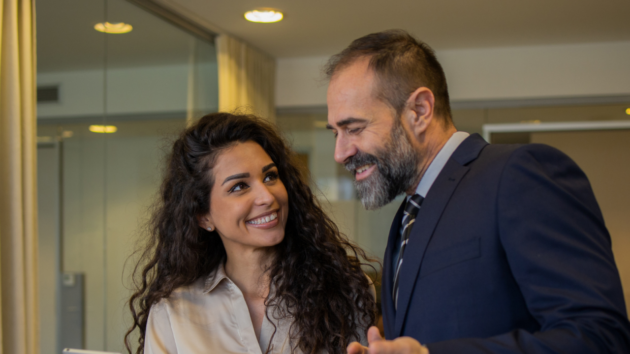 Two professionals in a modern office setting are engaged in a discussion. One person is holding a tablet while the other gestures with their hands, indicating collaboration and decision-making related to office operations. The background includes glass walls and a conference area, emphasizing a corporate environment.