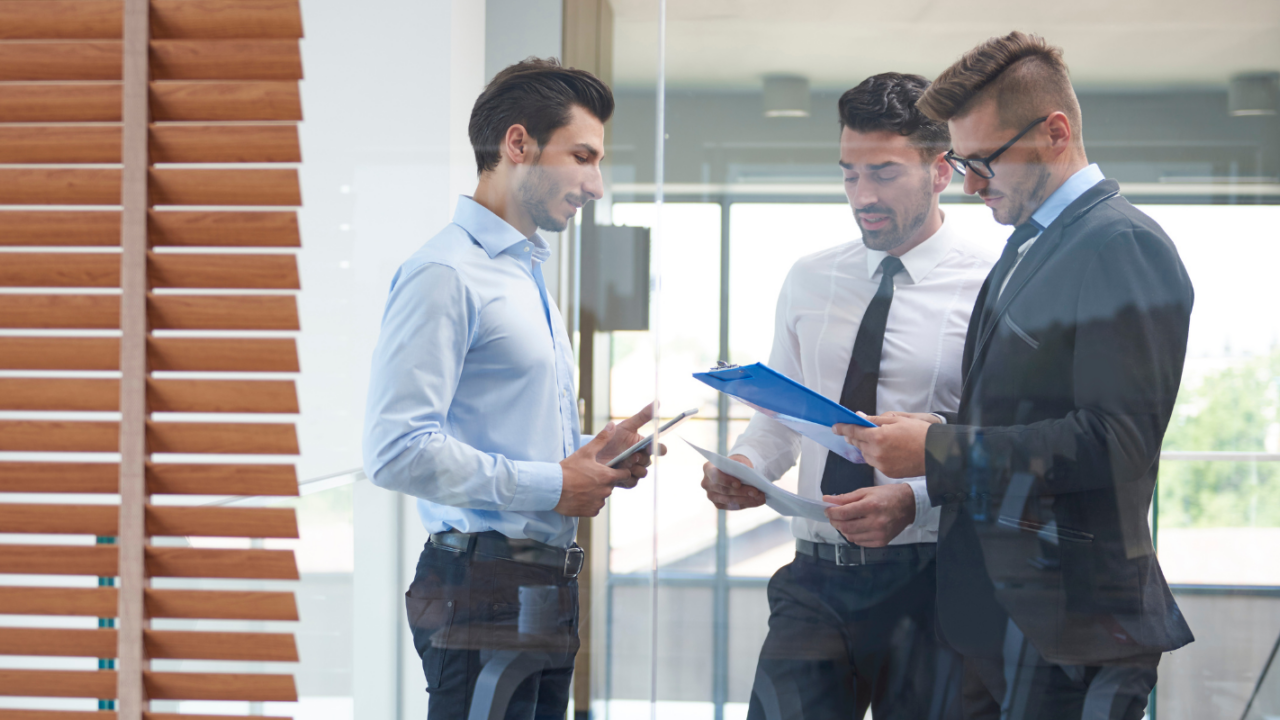 Three professionals stand in a modern office reviewing financial reports on a tablet and clipboard, discussing budgeting, forecasting and financial performance as part of financial planning and analysis.