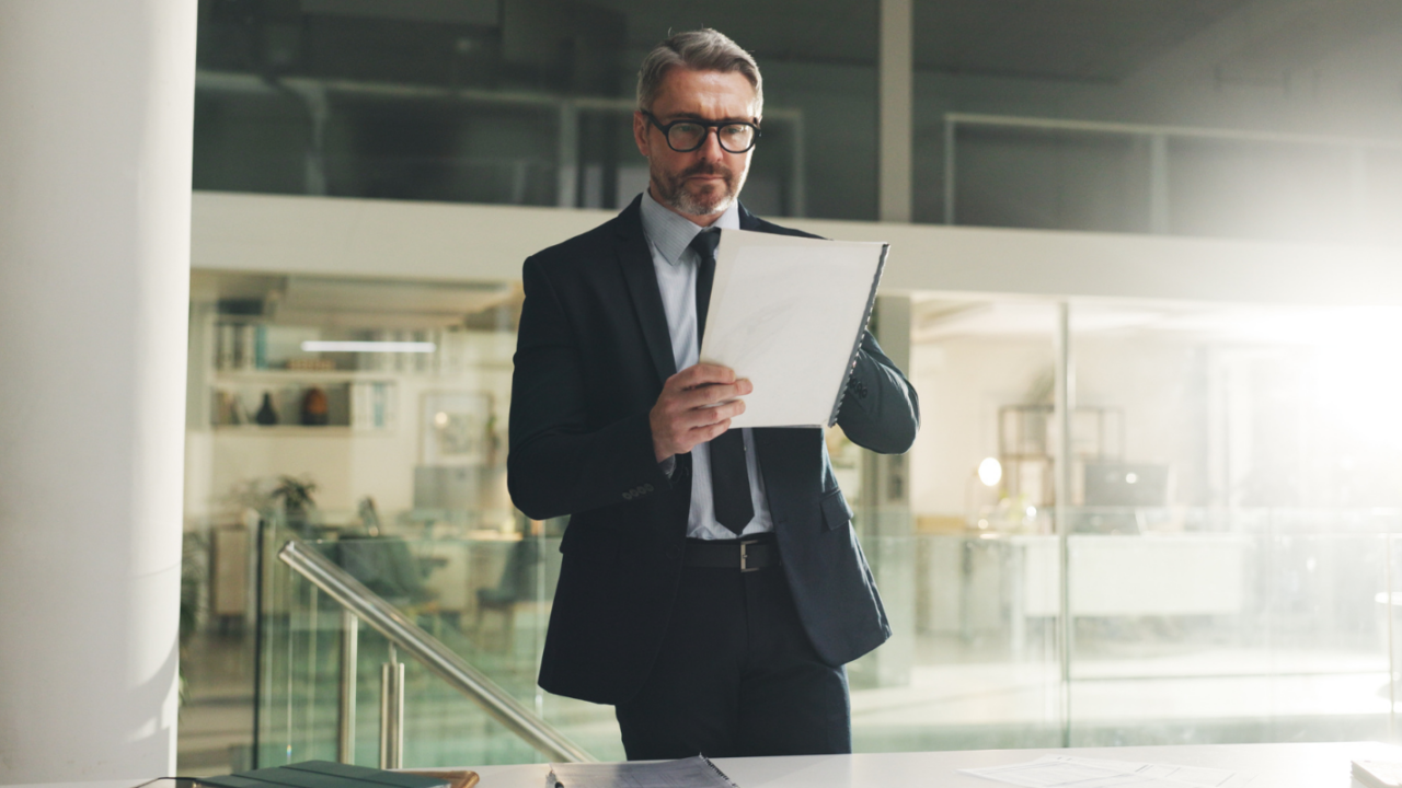 Person in a formal business suit standing in a modern office environment, holding and reviewing documents related to executive and interim leadership.