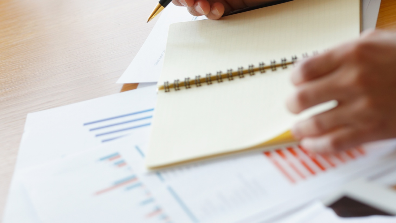 Close-up of a person writing in a notebook while reviewing financial charts and documents related to treasury and cash management on a wooden desk.