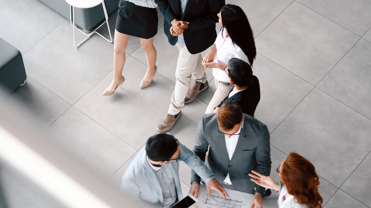 A group of professionals standing in a modern office space, gathered around a document and tablet, discussing plans and collaborating on a proposal.