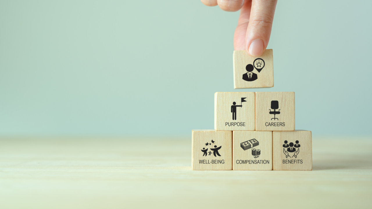 Hand placing a wooden block labeled leadership on top of a stack representing well being, compensation, benefits, purpose, and careers