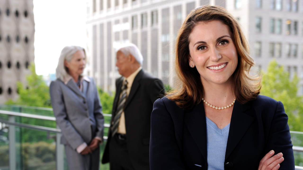 A human resources generalist standing confidently on an outdoor office terrace, representing corporate HR support and employee relations, with two colleagues conversing in the background.
