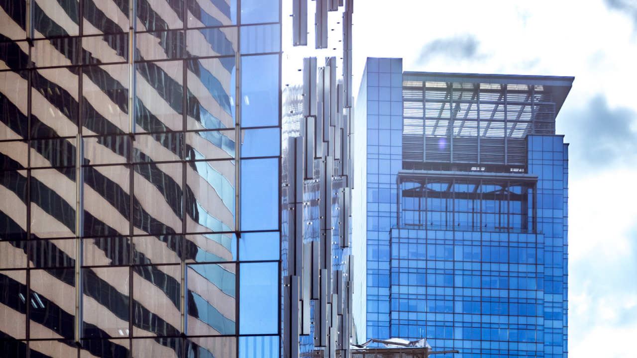 Tall modern office buildings with reflective glass facades in an urban business district. The structures feature sleek architectural designs with blue-tinted windows and metallic accents, symbolizing corporate real estate and commercial property development.