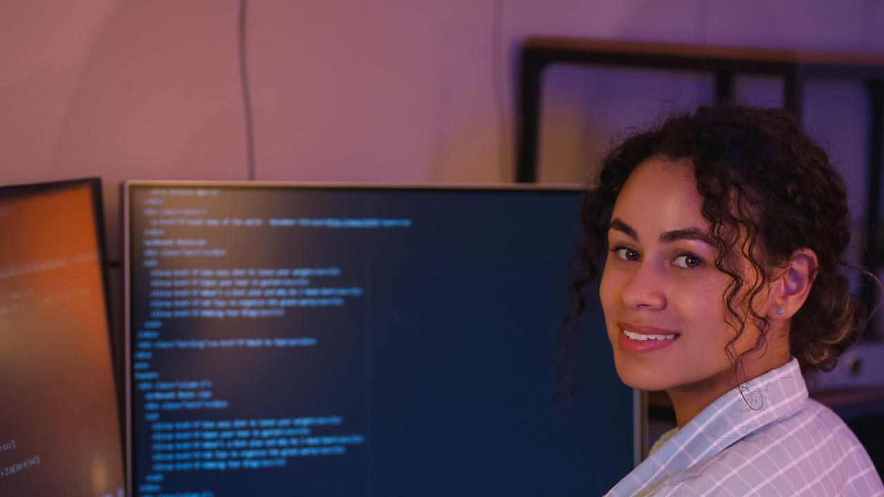 Person seated at a desk with two large monitors displaying lines of code and system scripts in a dimly lit environment, representing tech operations and system monitoring tasks.