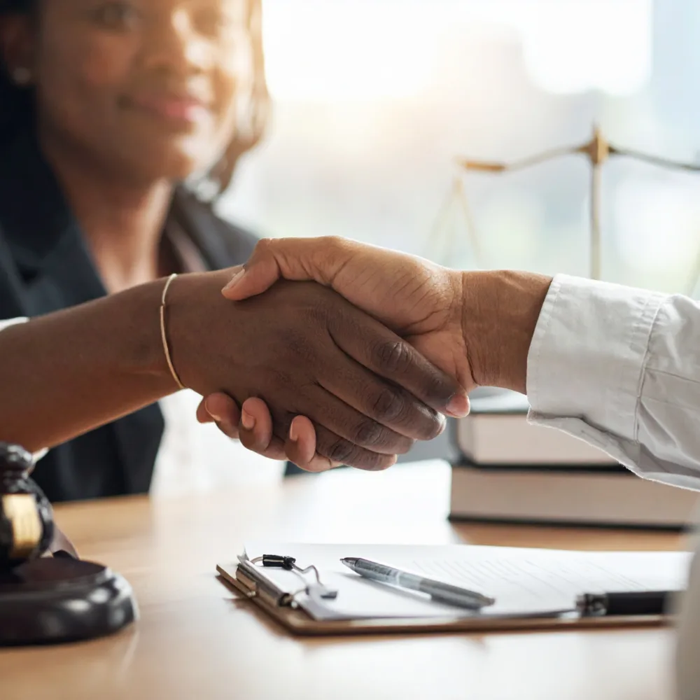 Two legal professionals shaking hands across a desk with case documents and a clipboard, symbolizing a formal legal agreement or client consultation.