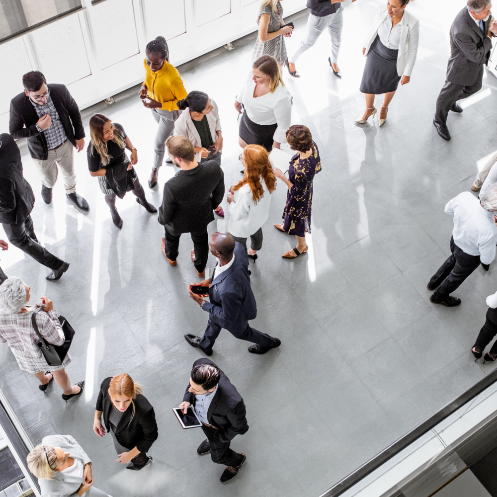 A large group of professionals gathered in a bright, modern indoor space, engaging in conversations and networking. The setting includes people standing in small clusters, some holding drinks or tablets, representing a corporate event or business gathering.