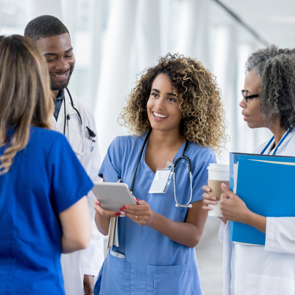 A group of medical professionals in scrubs and lab coats standing in a bright hospital corridor, engaged in conversation. One person holds a tablet, another carries a clipboard, and a third has a coffee cup, symbolizing collaboration and teamwork in healthcare settings.