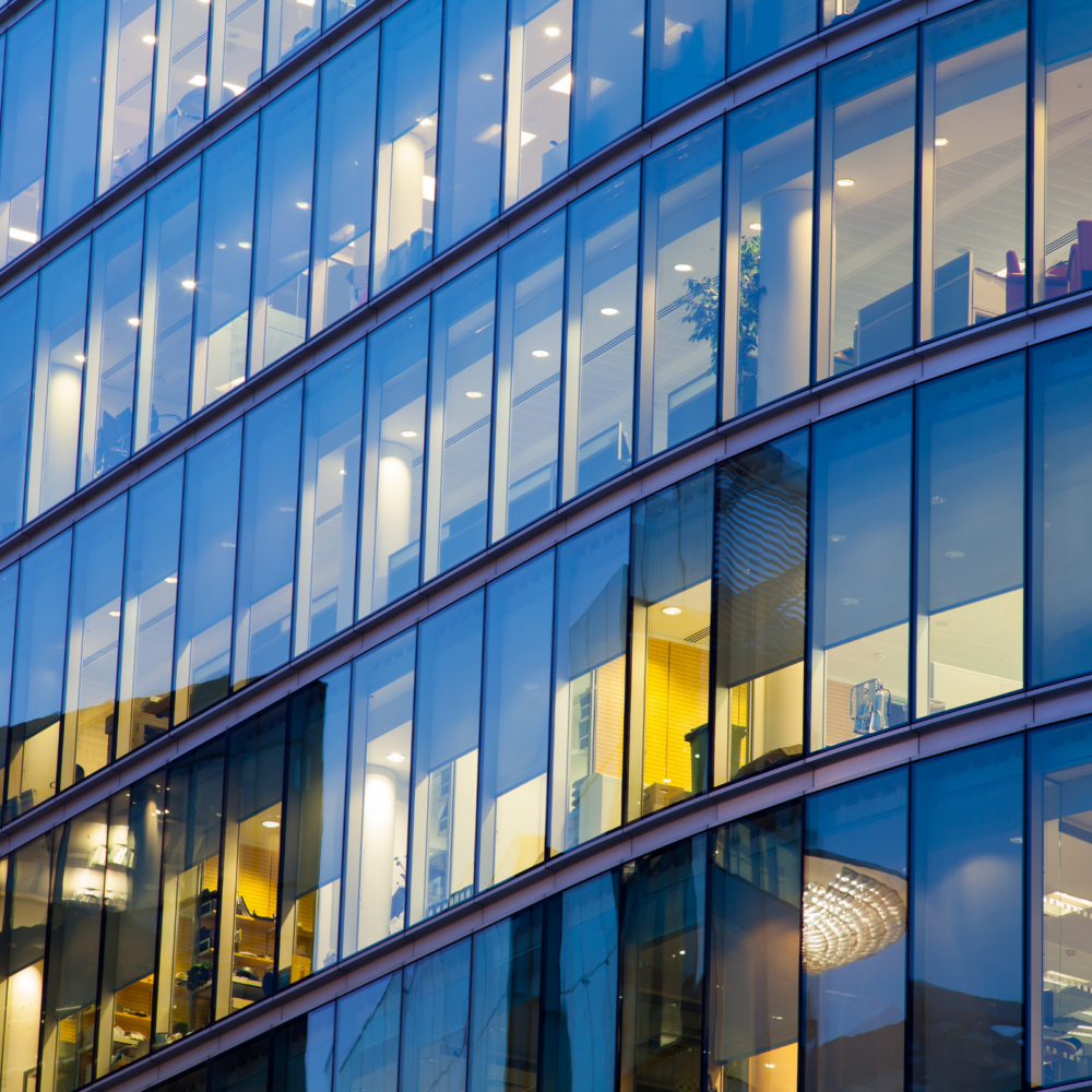 Exterior view of a contemporary office building with large glass windows revealing multiple floors. Inside, well-lit workspaces, meeting areas, and modern furnishings are visible, showcasing a professional and collaborative office environment.