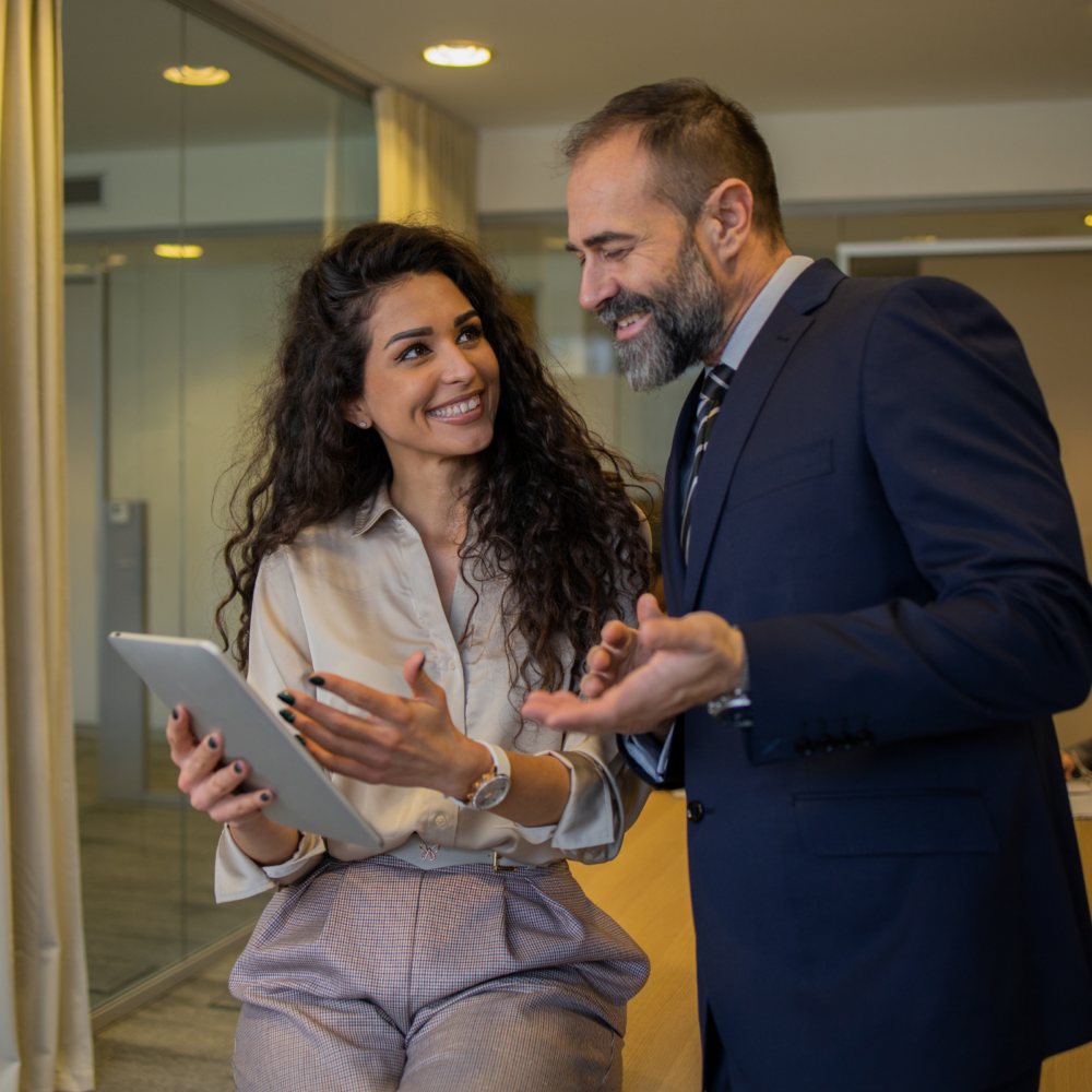 Two professionals in a modern office setting are engaged in a discussion. One person is holding a tablet while the other gestures with their hands, indicating collaboration and decision-making related to office operations. The background includes glass walls and a conference area, emphasizing a corporate environment.
