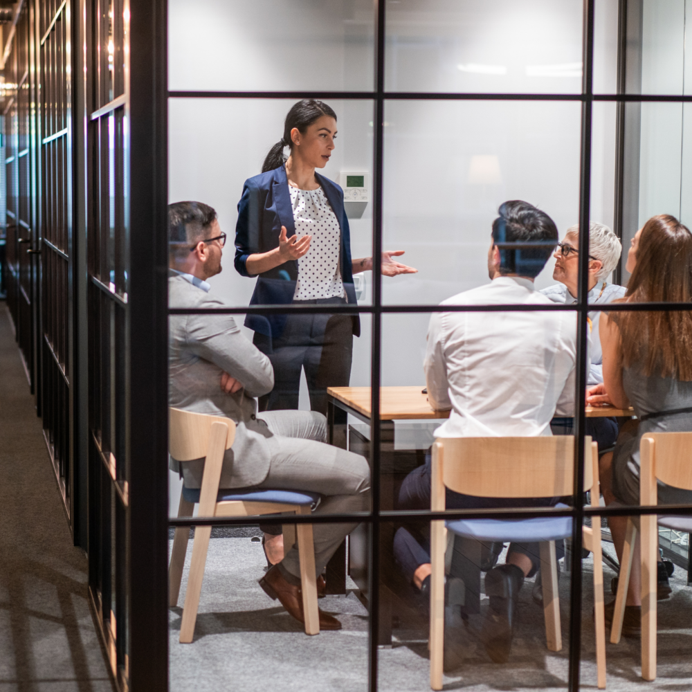 A modern glass-walled conference room with several professionals seated around a wooden table while another person stands and speaks, gesturing during a presentation. Laptops and documents are visible on the table, representing collaboration and strategy planning for sales and business development.