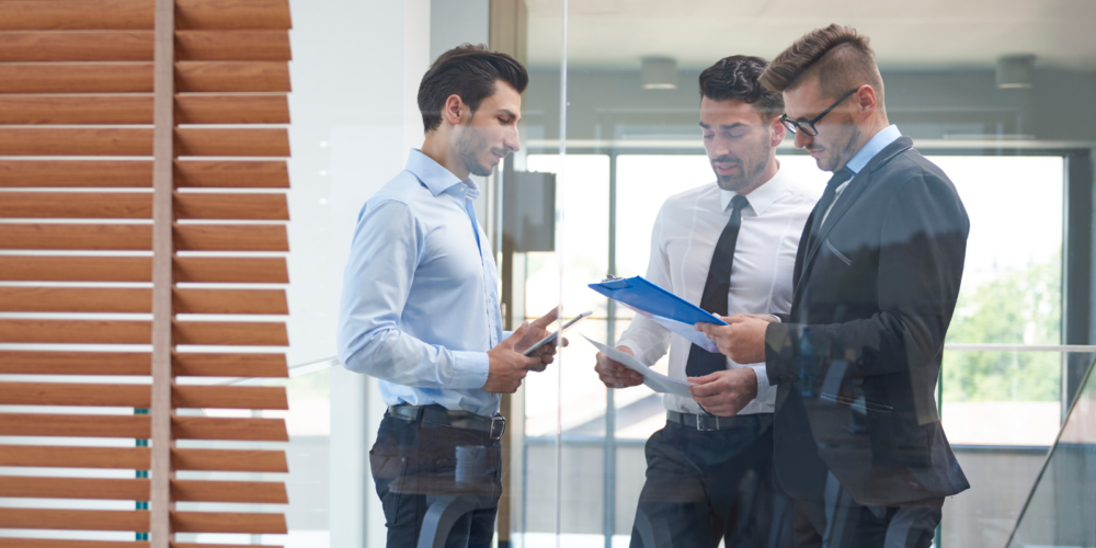 Three professionals stand in a modern office reviewing financial reports on a tablet and clipboard, discussing budgeting, forecasting and financial performance as part of financial planning and analysis.