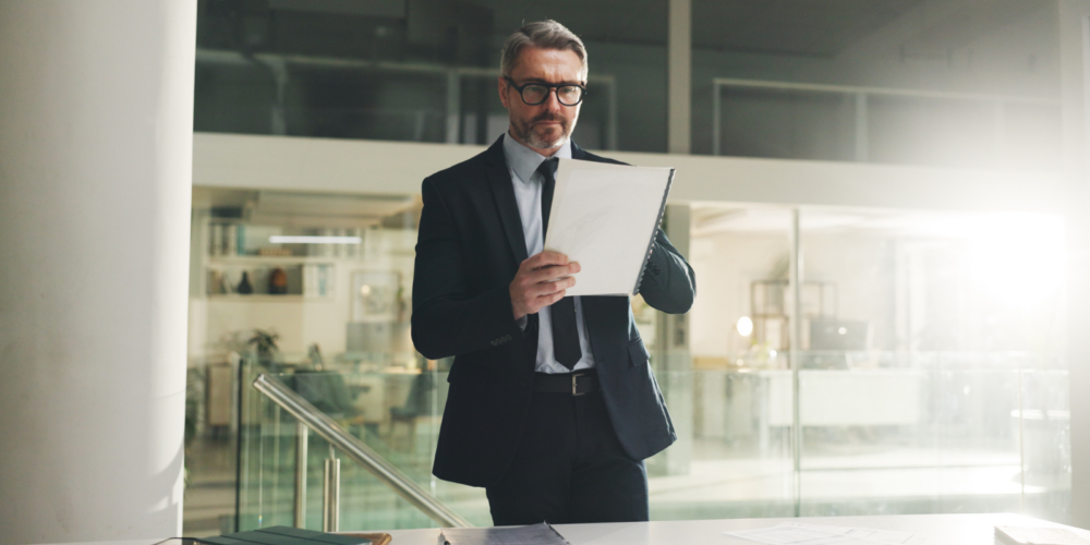 Person in a formal business suit standing in a modern office environment, holding and reviewing documents related to executive and interim leadership.
