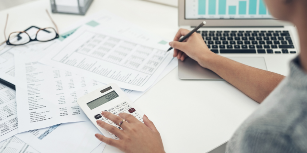 Person reviewing payroll spreadsheets and financial data using a calculator and laptop with charts on the screen.