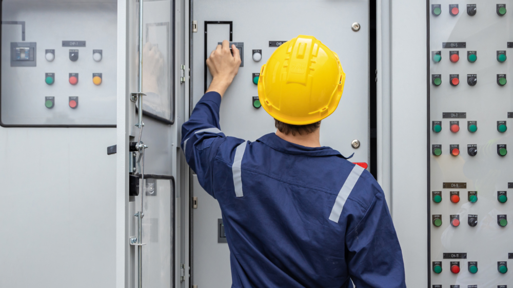 An engineer wearing a yellow safety helmet and blue protective coveralls operates a control panel in an industrial setting. The panel features multiple switches, buttons, and indicators, suggesting work related to electrical systems or equipment maintenance.