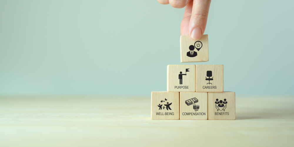 Hand placing a wooden block labeled leadership on top of a stack representing well being, compensation, benefits, purpose, and careers