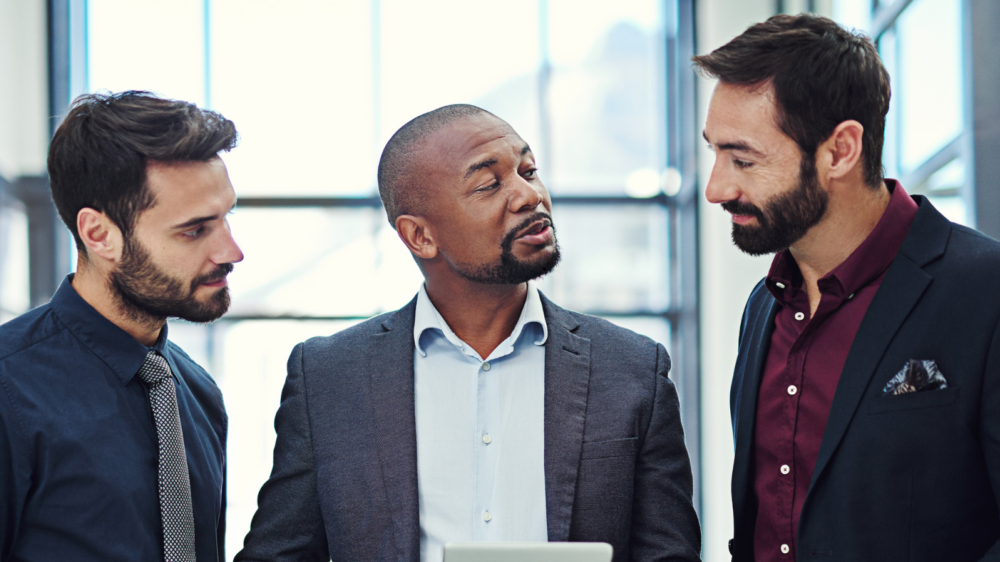 A human resources business partner holding a tablet speaks with two colleagues in a modern office setting, engaged in a collaborative discussion.